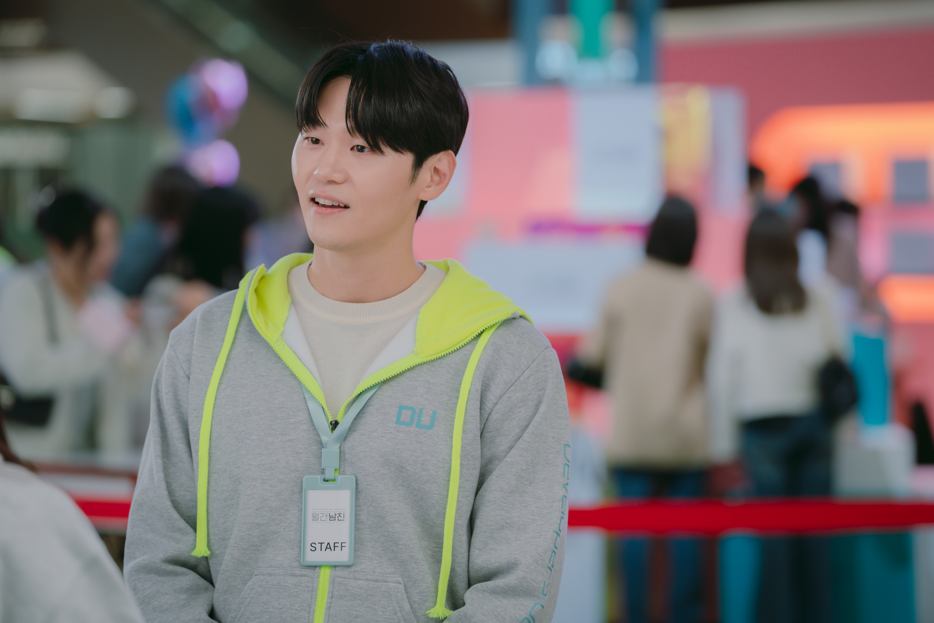 A man (Lee Hak-joo as Min Jun-young) wears a gray hoodie with neon lining, while standing in front of a crowded mall pop-up, in the Netflix K-drama 'Boyfriend on Demand.'