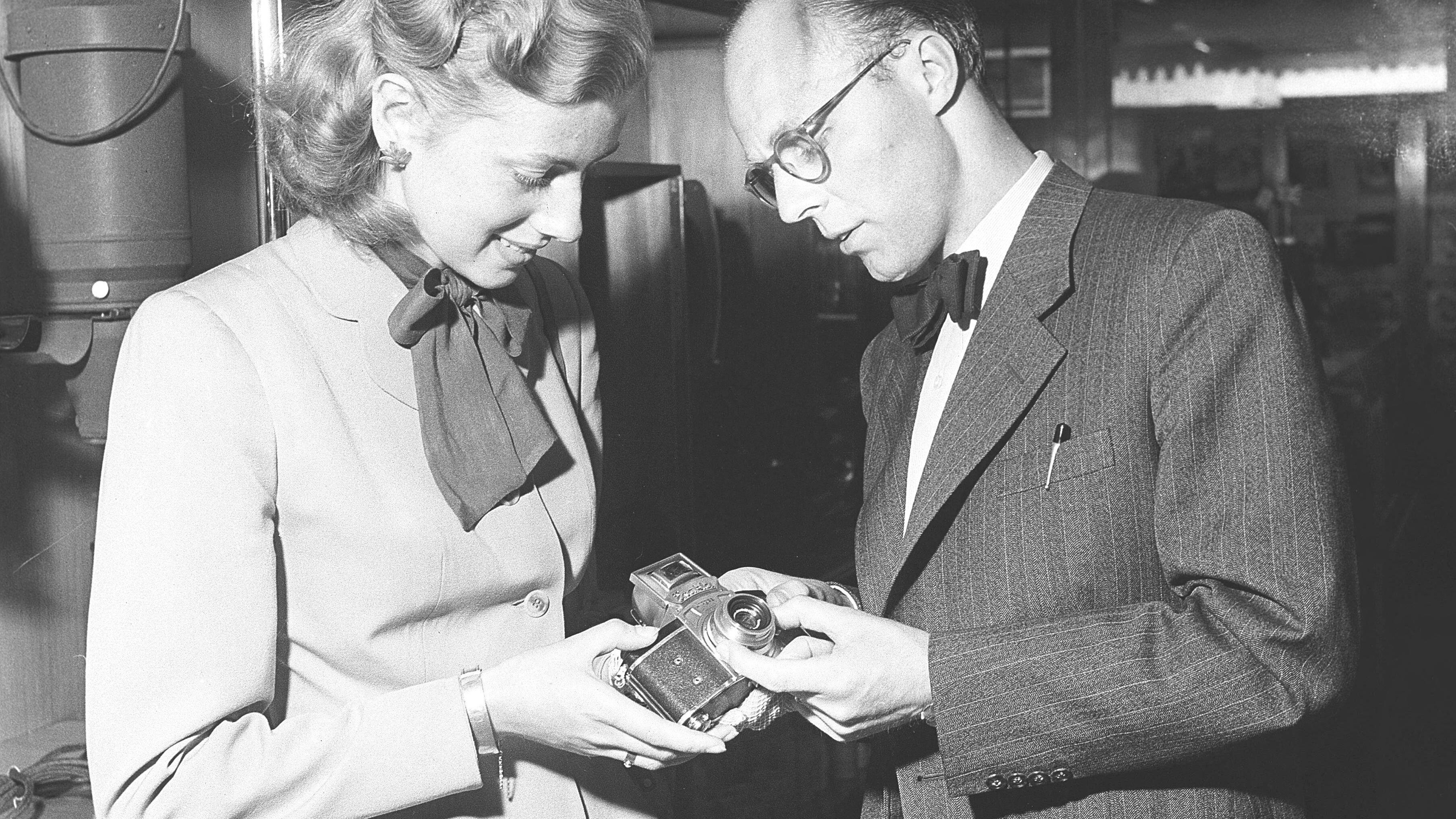 3CKHWC9 In a camera and photography equipment store in 1949, a young woman is shown the camera Exakta II. The Exakta II, introduced in 1949 by Ihagee Kamerawerk in Dresden, was a compact 35mm single-lens reflex (SLR) camera, building on the pre-war Kine Exakta design. Produced in limited quantities under Soviet control post-World War II, it featured a left-sided shutter release, waist-level viewfinder, and compatibility with premium Zeiss Jena and Meyer lenses. That year, a pentaprism accessory enabled eye-level viewing, boosting its appeal. Marked Made in Germany, it became a sought-after import in