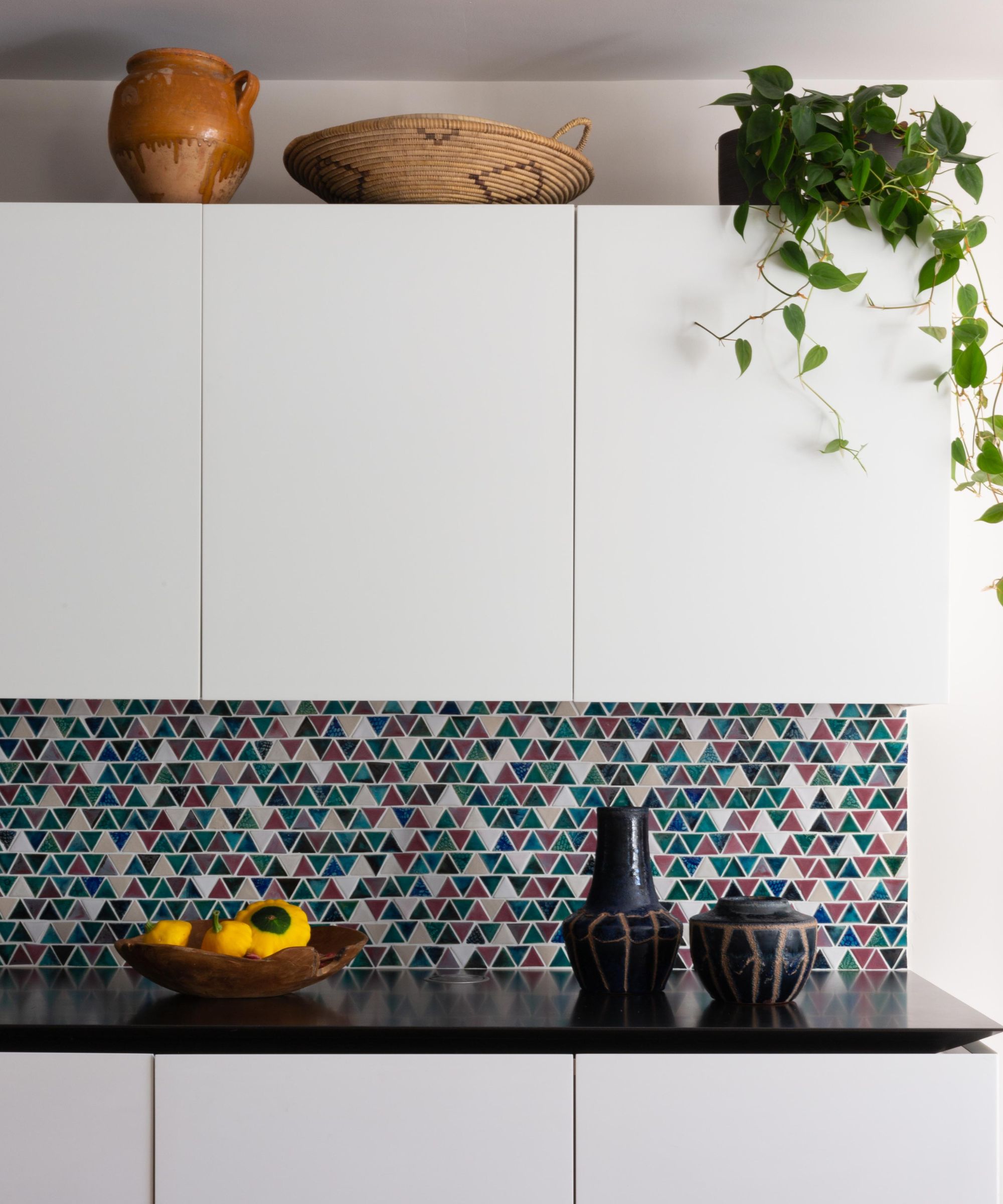 A minimal white handleless kitchen with a striking multicoloured triangular mosaic tile splashback in teal, plum and cream. A black worktop is styled with ceramic vases and a wooden bowl of yellow fruit, while a trailing plant, terracotta jug and woven basket sit on top of the upper cabinets.