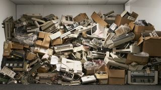 A huge pile of legacy technology sitting in an old-fashioned office environment, with old printers, CRT monitors, keyboards, and cardboard boxes piled up in a stack of trash taller than a person.