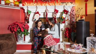 Two women with a dog in a Christmas jumper with lots of stockings behind them