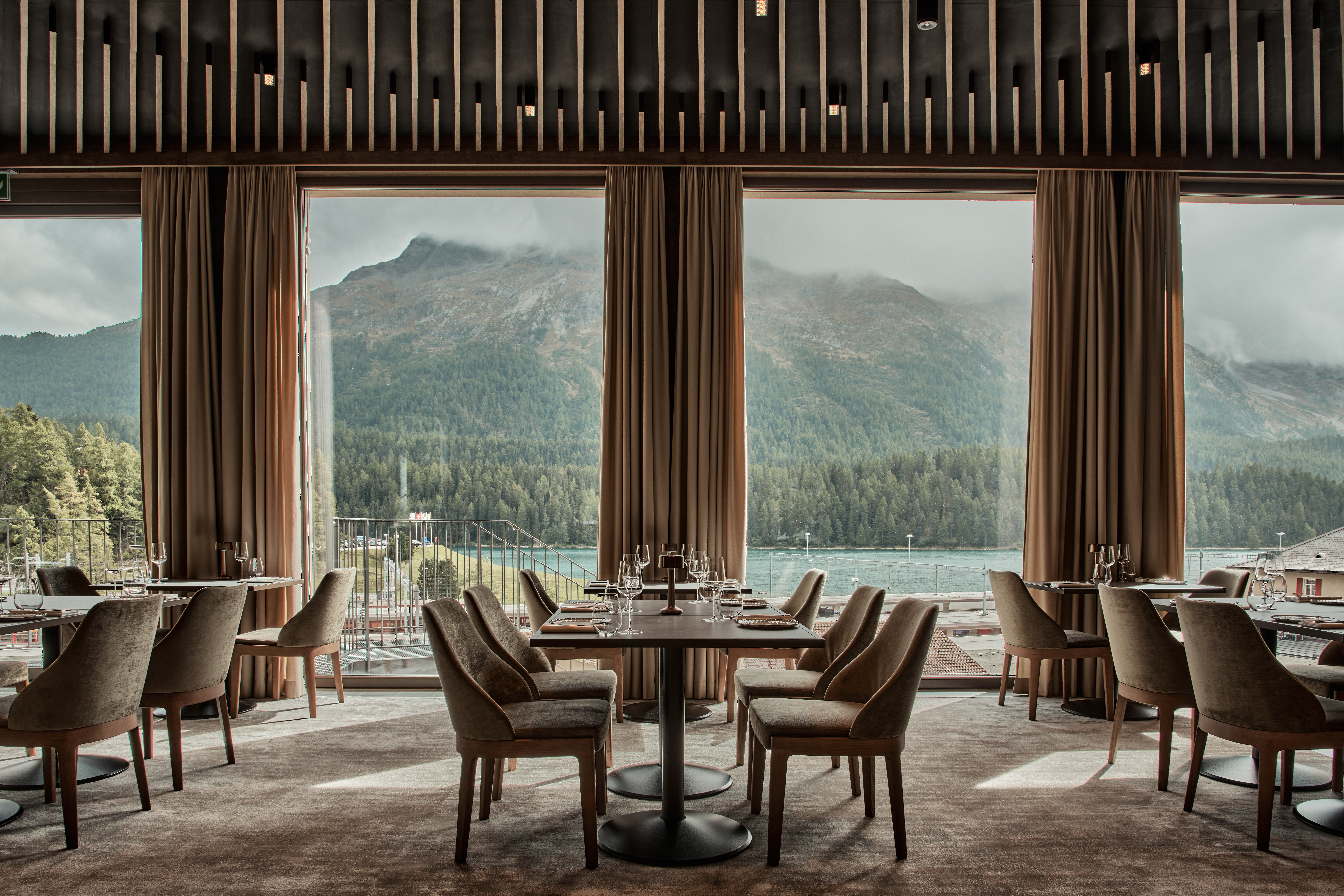 A panoramic hotel restaurant boasts floor-to-ceiling windows punctuated by beige, flowy curtains that open up to a misty lakeview.