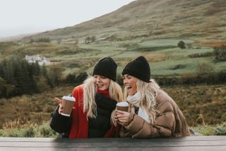 Two girls drinking a coffee at a picnic bench in a rural setting