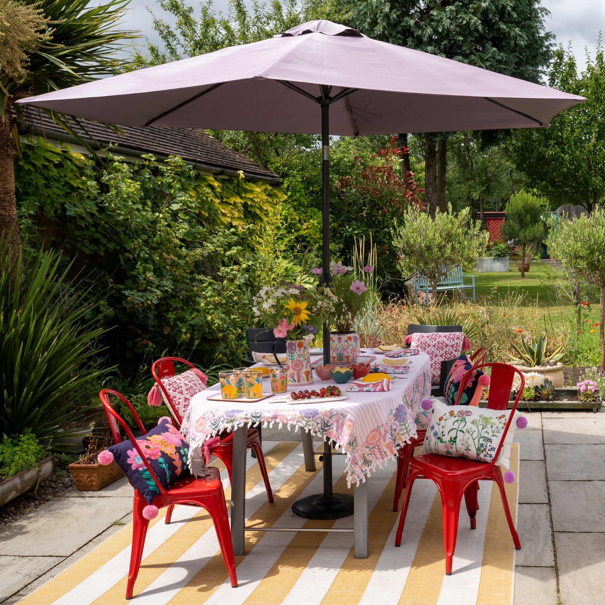 A garden with a colourfully set outdoor dining table with red metal chairs and a parasol