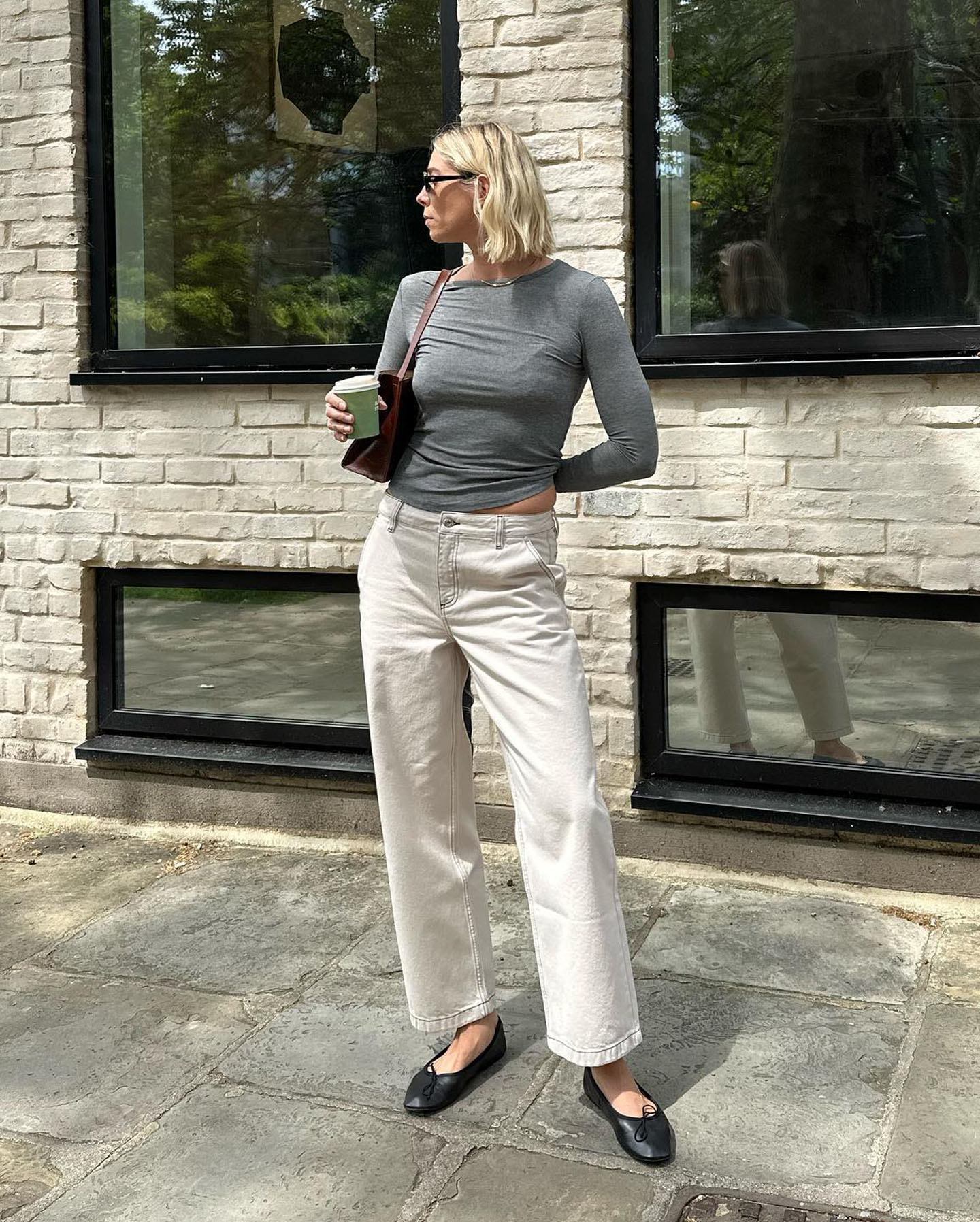 stylish influencer Lindsey Holland poses with her short blond bob on a sunny sidewalk wearing a long sleeve gray t-shirt, brown shoulder bag, off-white jeans, and black ballet flats