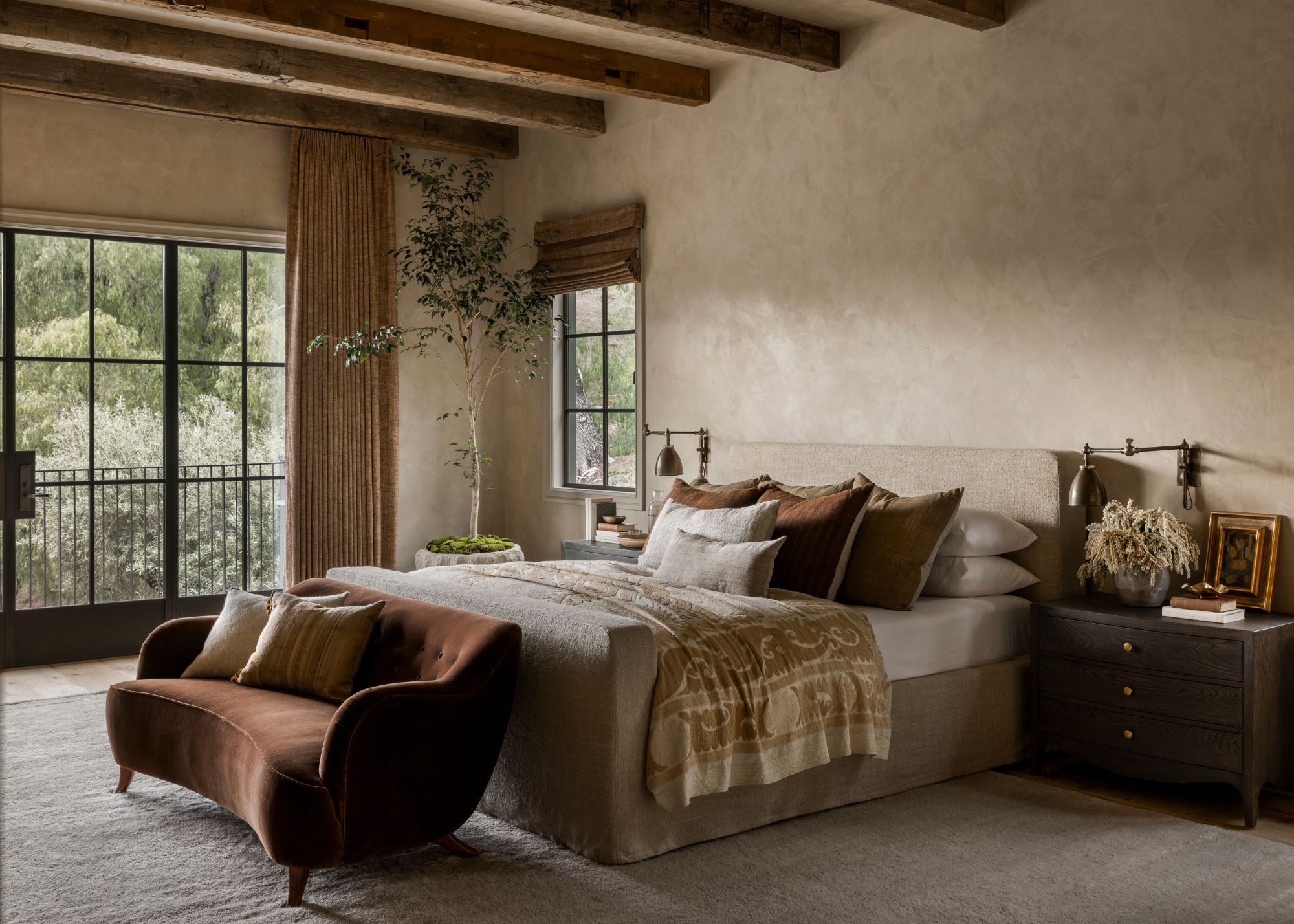 A large modern neutral bedroom with exposed wooden beams on the ceiling, a small rust sofa at the foot of the bed, a large potted tree in the corner of the room and black bedside tables