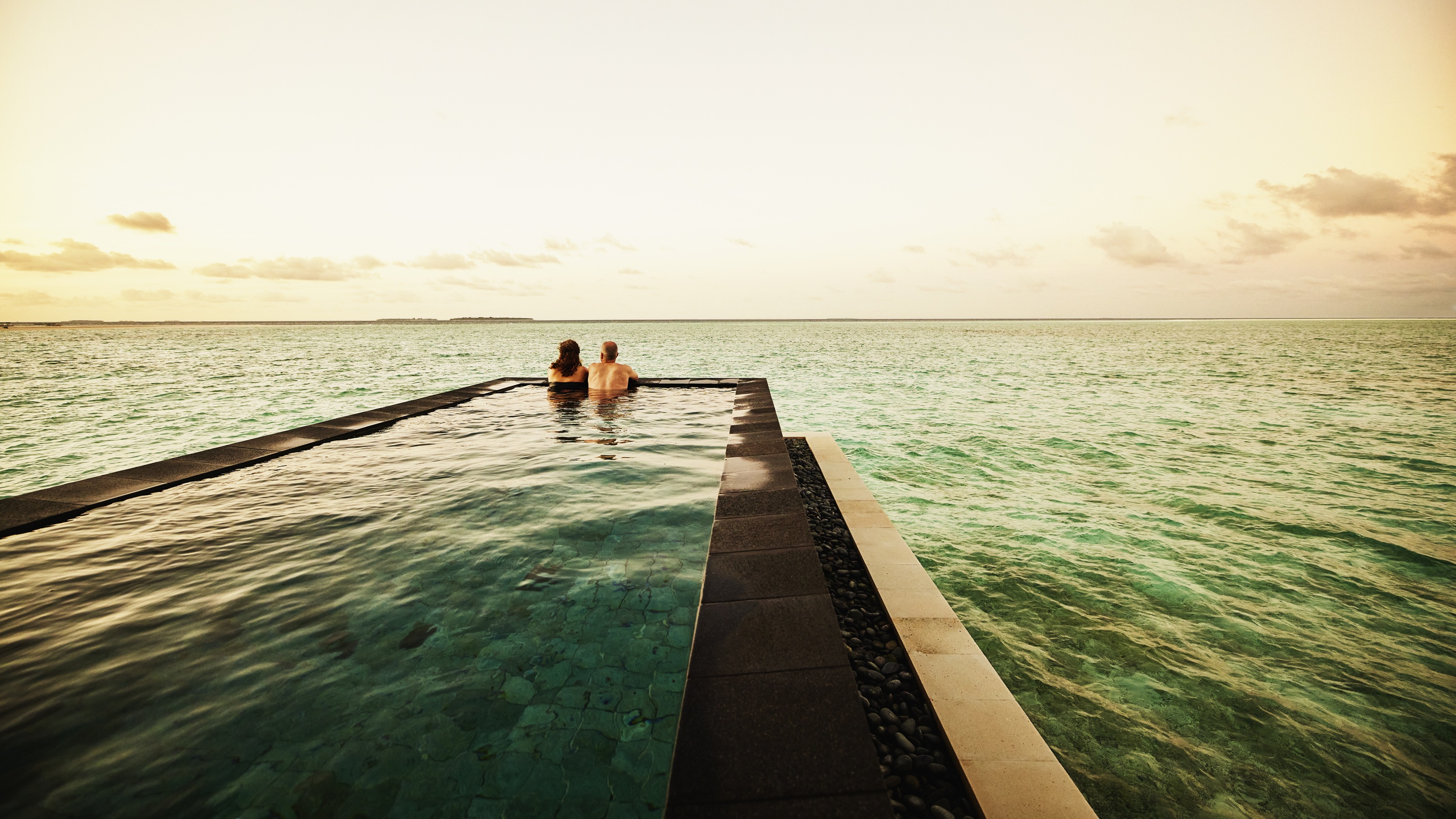 A couple relax in an infinity pool while looking out at the sea at sunset.
