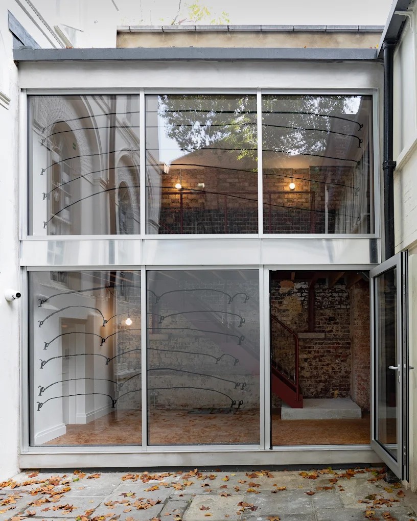 The glass facade of a gallery space, whose outdoor patio is covered in brown leaves.