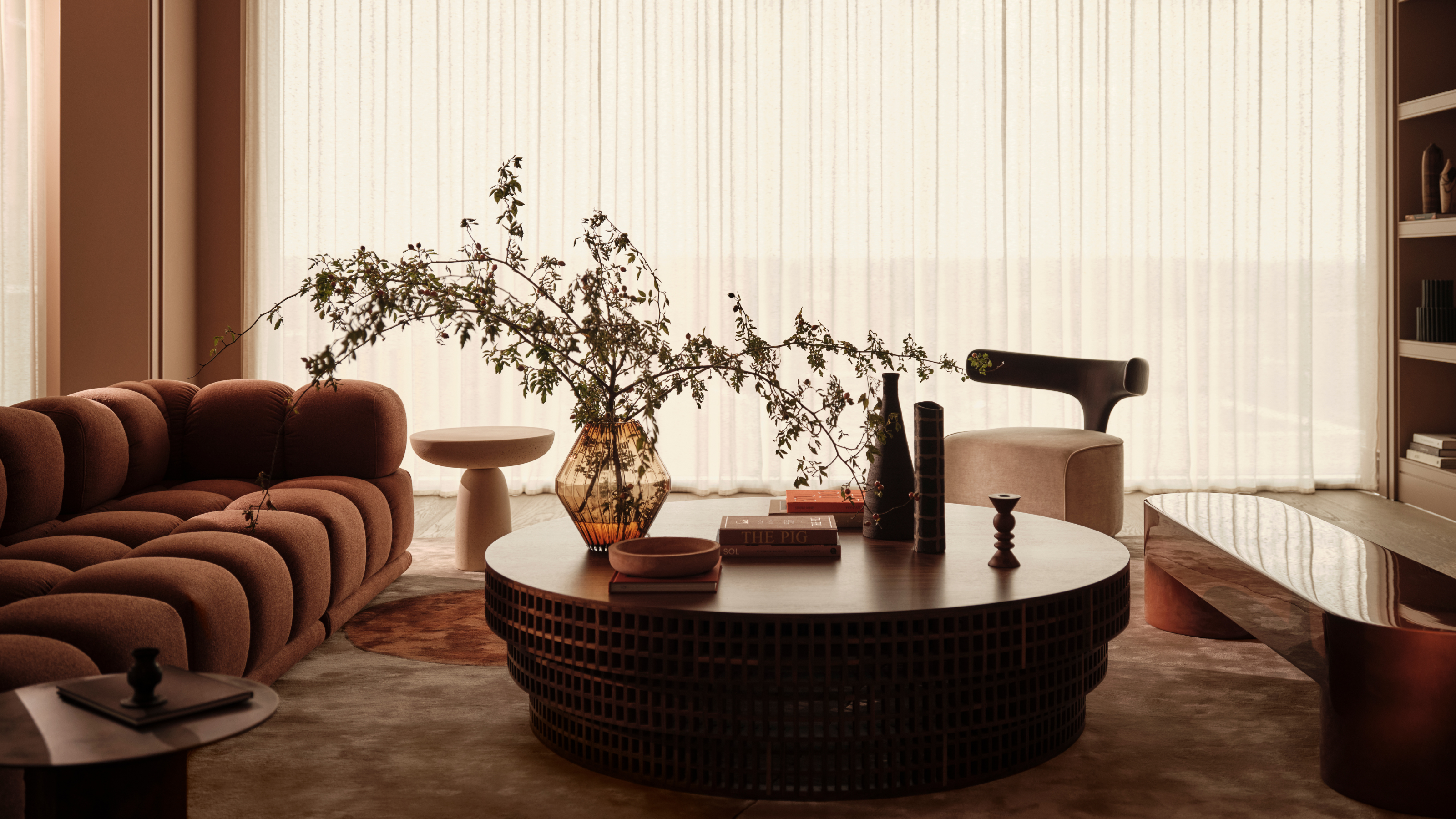 A modern living space with a rust sofa, a large round coffee table in the center of the space with foliage in a vase on top and closed sheer curtains across a large floor to ceiling window