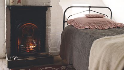 Cozy white bedroom with exposed bricks, fireplace with lit fire, and black bed with grey bedding, white blanket, and white and red striped scalloped pillows