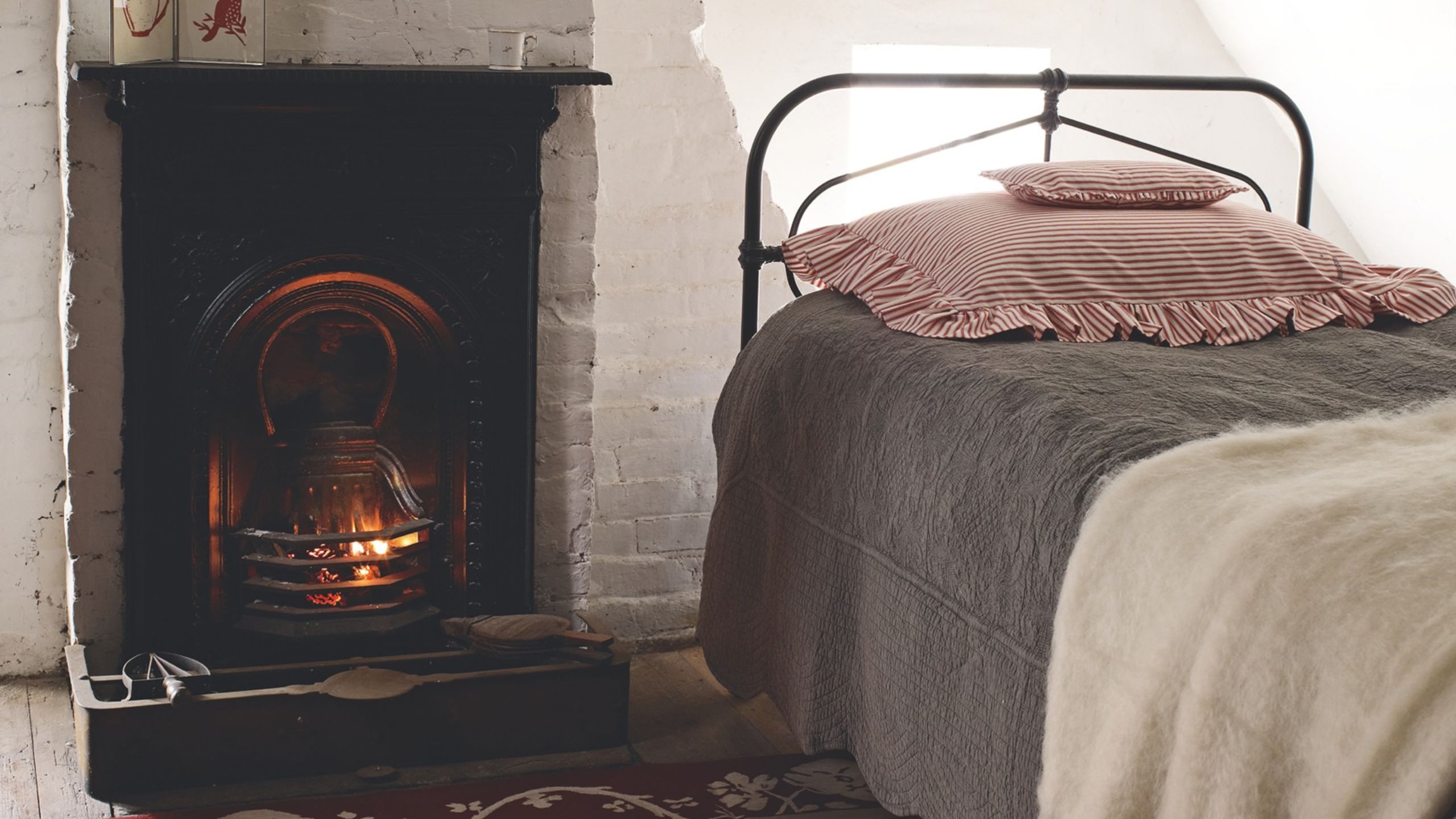 Cozy white bedroom with exposed bricks, fireplace with lit fire, and black bed with grey bedding, white blanket, and white and red striped scalloped pillows