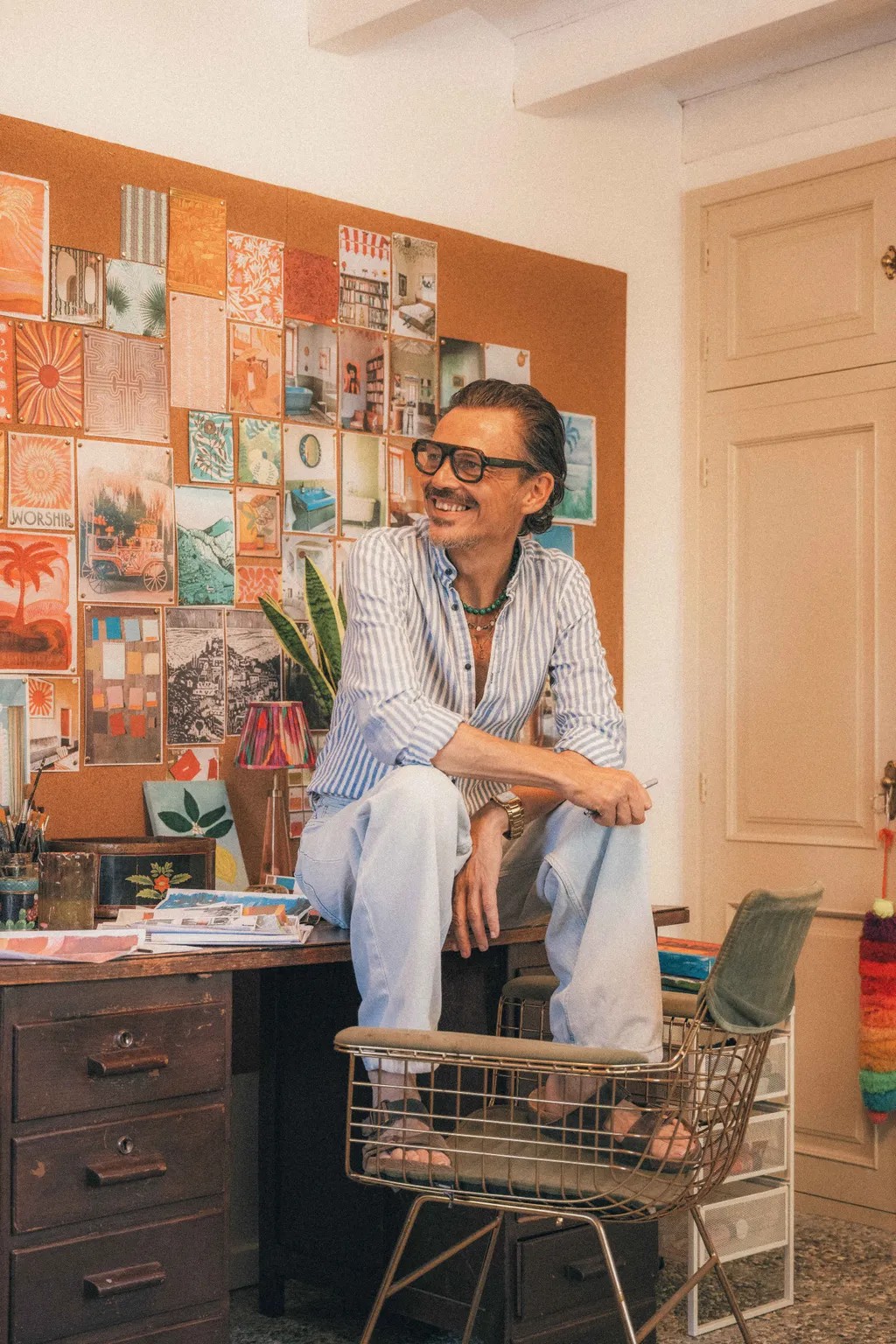 A middle-aged man in a stripy, blue and white shirt and pale denim jeans sit atop a busy desk in a colorfully decorated studio.