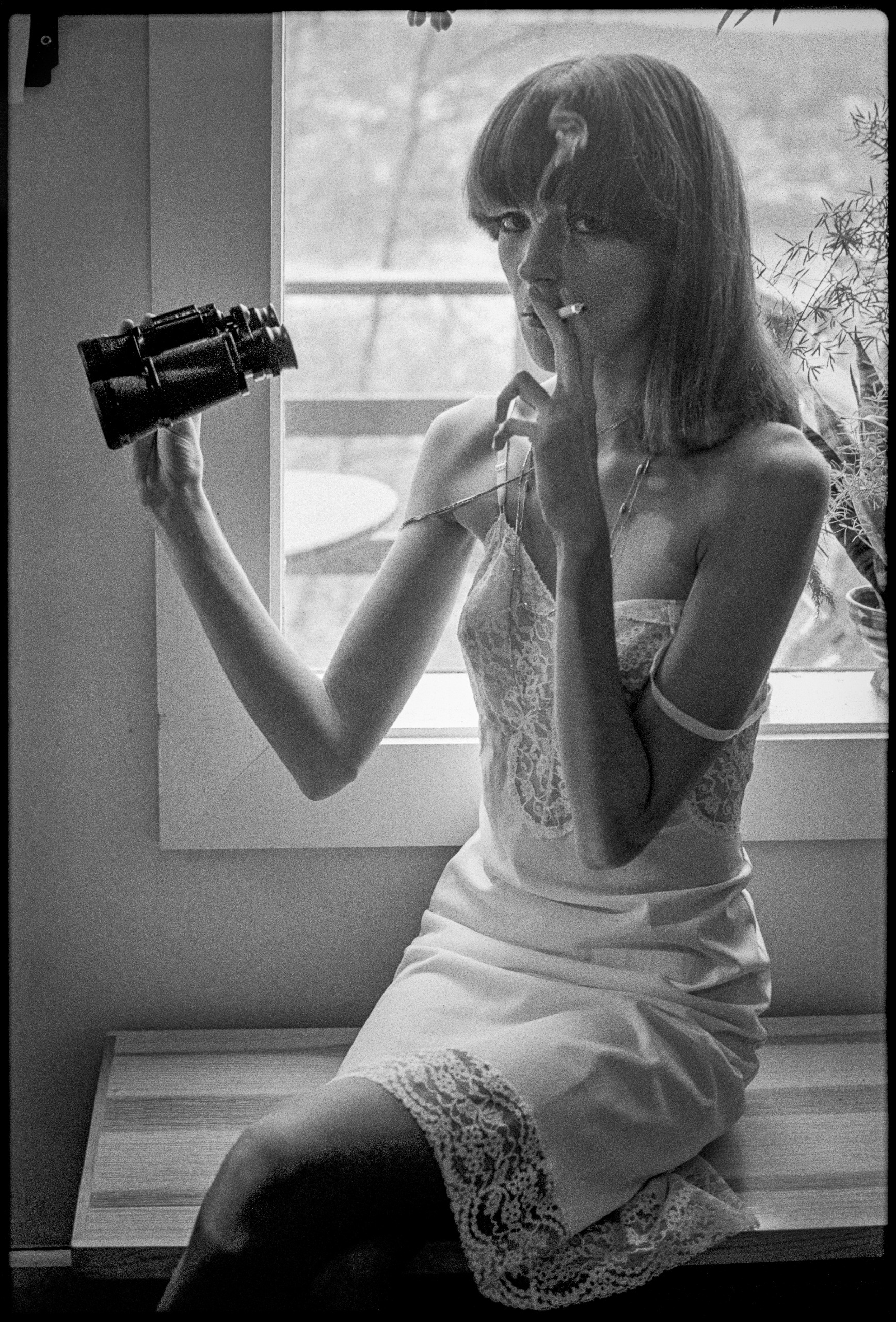 A black-and-white portrait of a woman sitting near a window in a lace trim slip dress, holding a pair of binoculars in one hand while smoking a cigarette.