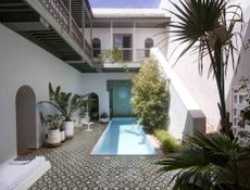 A hotel courtyard with a small long pool, loungers and patterned floor tiles next to a staircase.