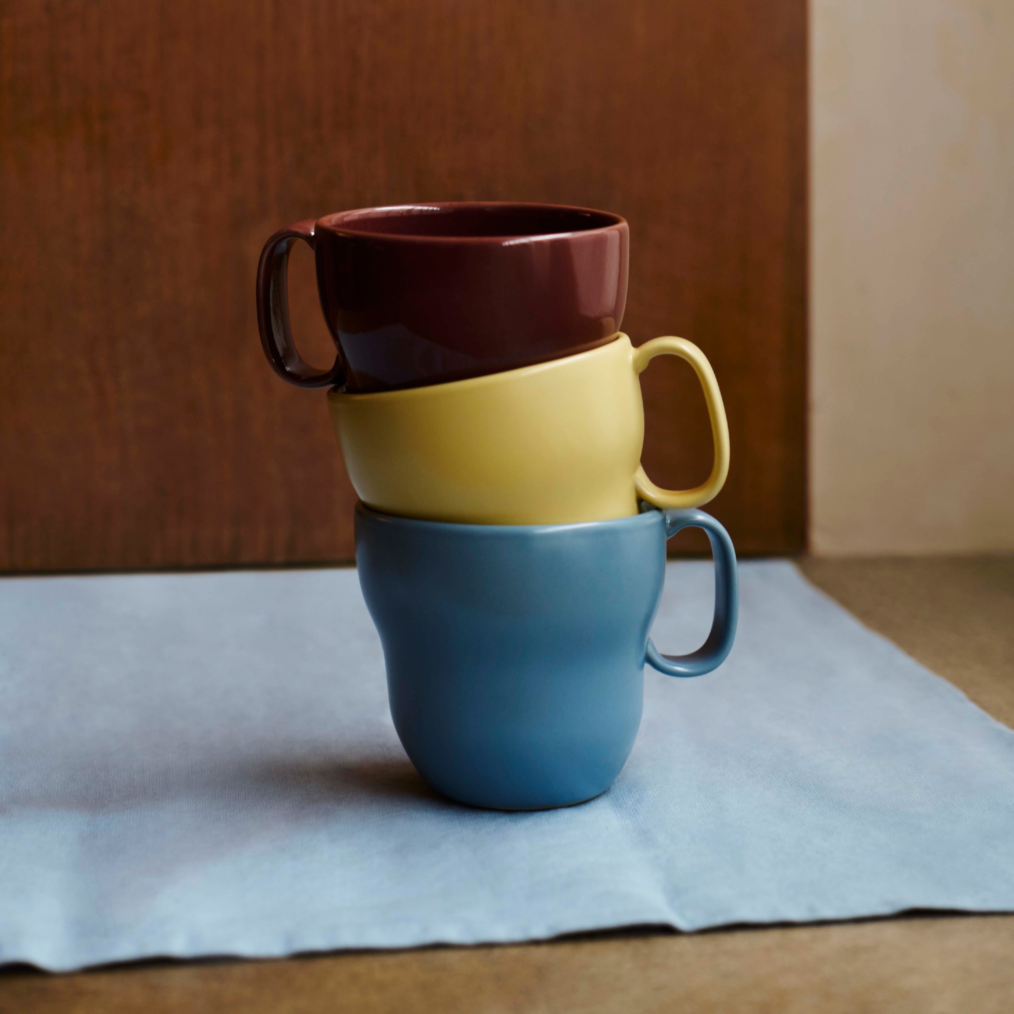 A stack of three mugs in light blue, butter yellow and burgundy on a light blue napkin