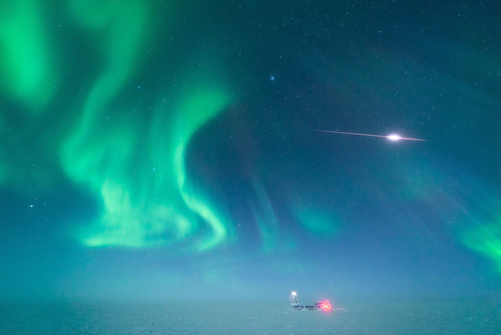 A bright meteor slicing the star-studded sky above the South Pole Telescope amid the shimmering lights of aurora australis.