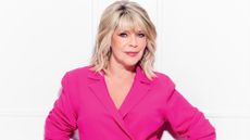 Ruth Langsford wears a hot pink suit and stands against a white panelled wall, looking glamorous