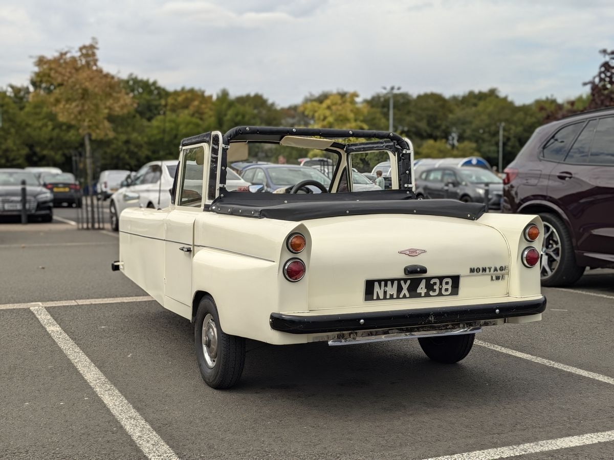 Cream-colored vintage three-wheeled convertible parked in a car park, viewed from the rear.