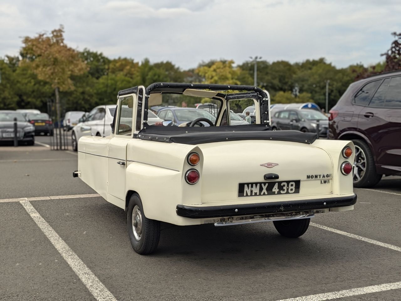 Cream-colored vintage three-wheeled convertible parked in a car park, viewed from the rear.