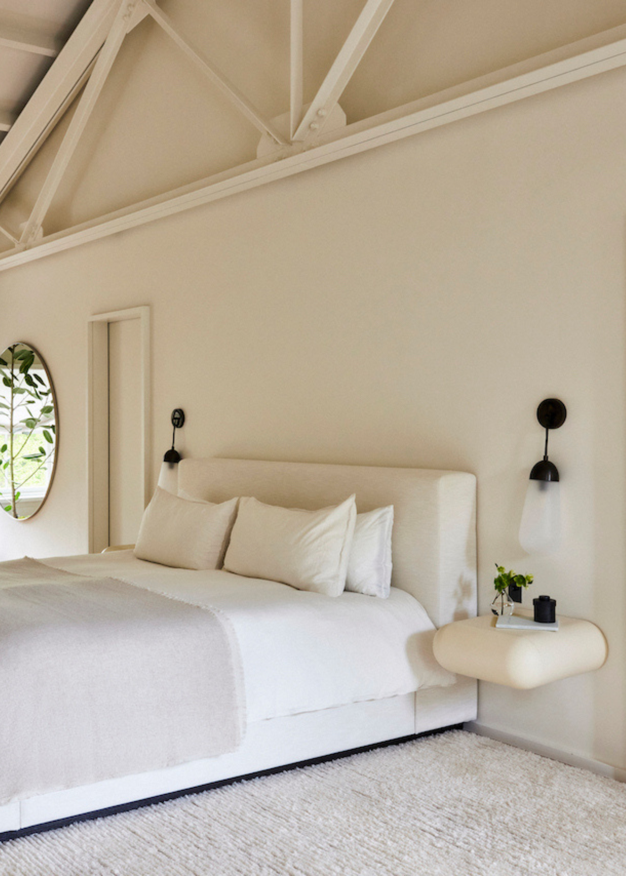 A white bedroom with tall ceilings, a white bed, a pair of black pendant wall sconces, a floating side table, and a white bedscape