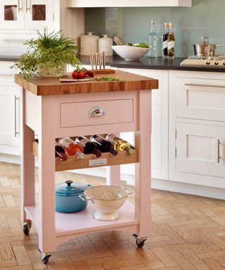 A pink freestanding kitchen island with wine bottles underneath it and white cabinets behind it