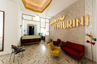 A quirkily decorated cafe and gallery space features terrazzo flooring in different colors, upholstered velvety furniture including armchairs and sofas, and colorful standing lamps.