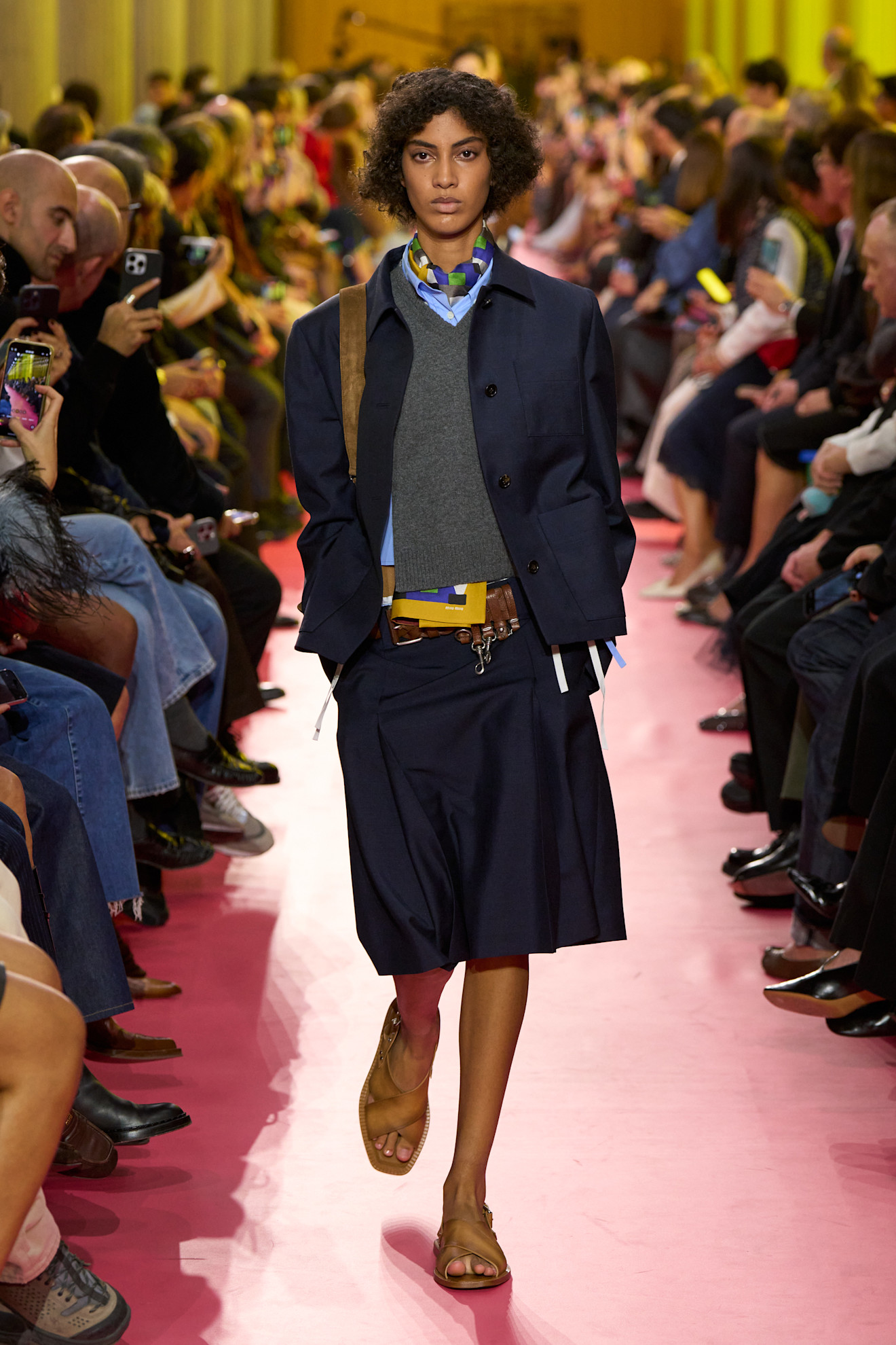 Layering staples for spring 2026 is shown in a photo from Miu Miu's runway show, with a model walking while wearing a navy blazer over a gray V-neck sweater and blue button-down shirt with a printed silk scarf tied around the neck, a navy midi skirt, tan slide sandals, and a tan handbag