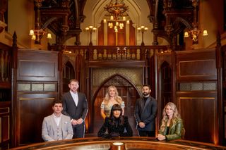 Claudia Winkleman sits at the roundtable in the Traitors castle with the Series 2 finalists, who are all dressed up smartly and formally. Sitting at the table on the left side of the picture is Harry Clark, with Andrew Jenkins standing behind him. Evie Morrison is standing behind Claudia, and Jaz Singh is standing next to her. Seated at the table on the right hand side of the picture is Mollie Pearce.