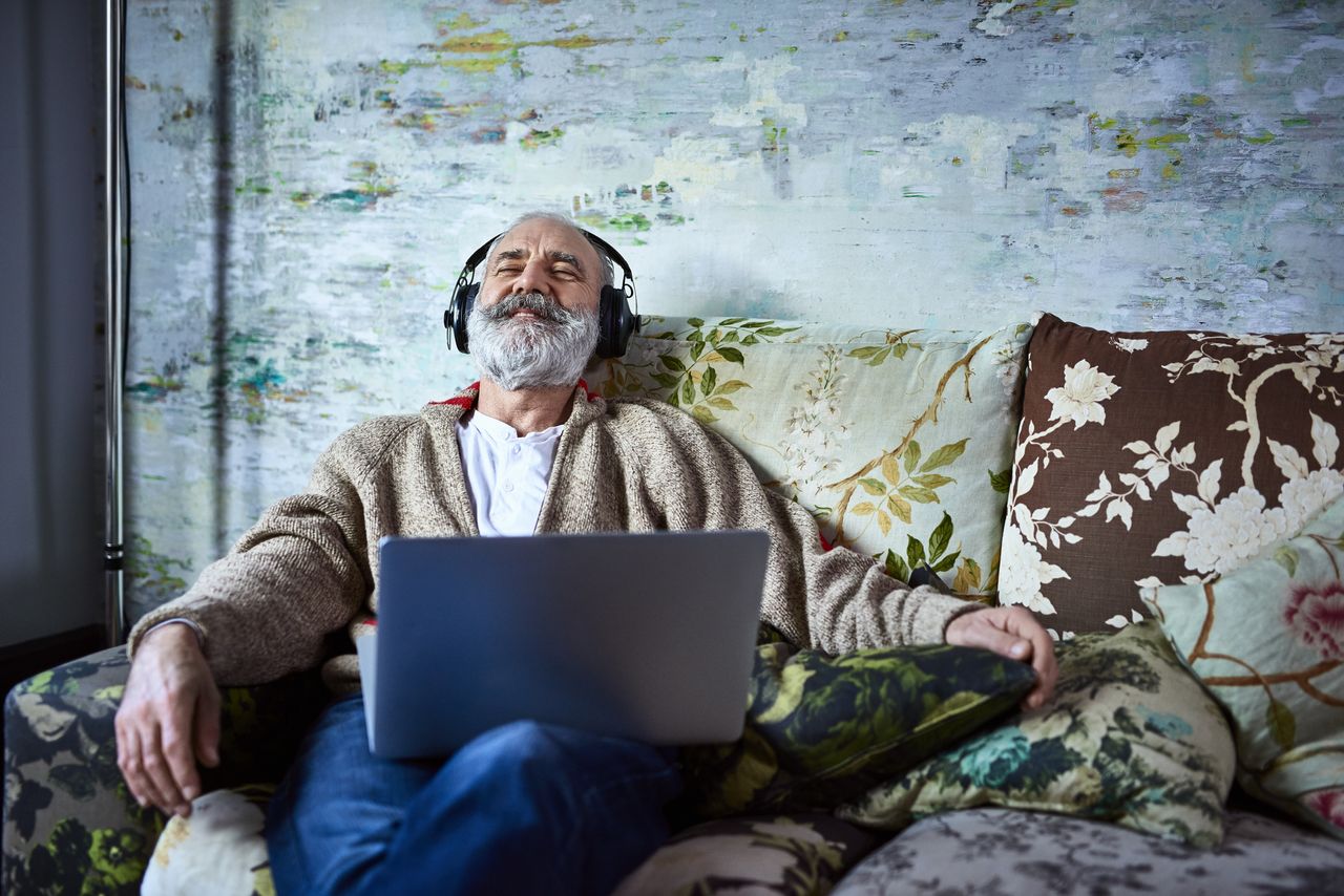 A man sits on a couch wearing headphones plugged into a computer and enjoys listening to music. 