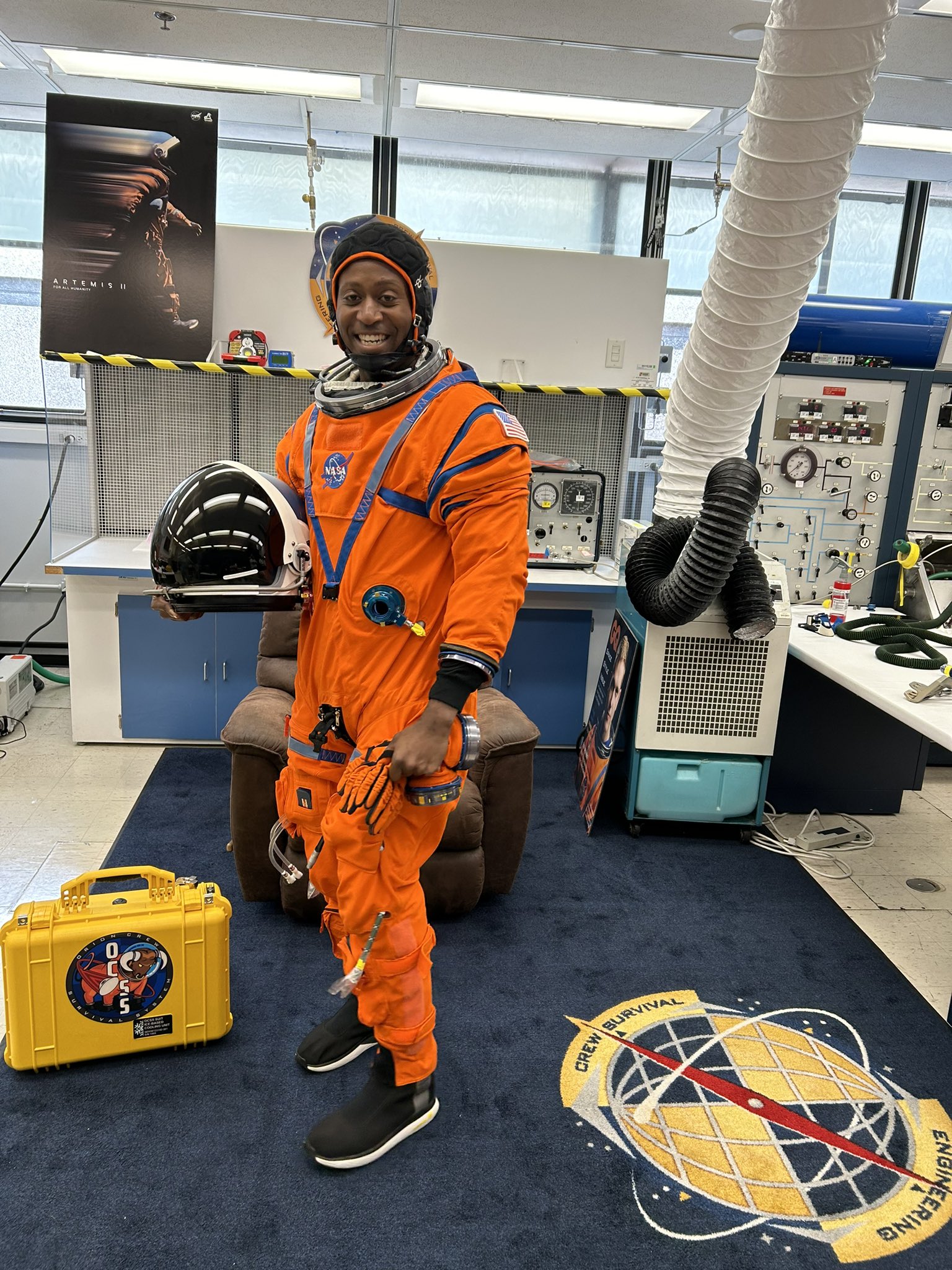 astronaut andre douglas smiling while standing up in an orange spacesuit, in an office filled with moon posters