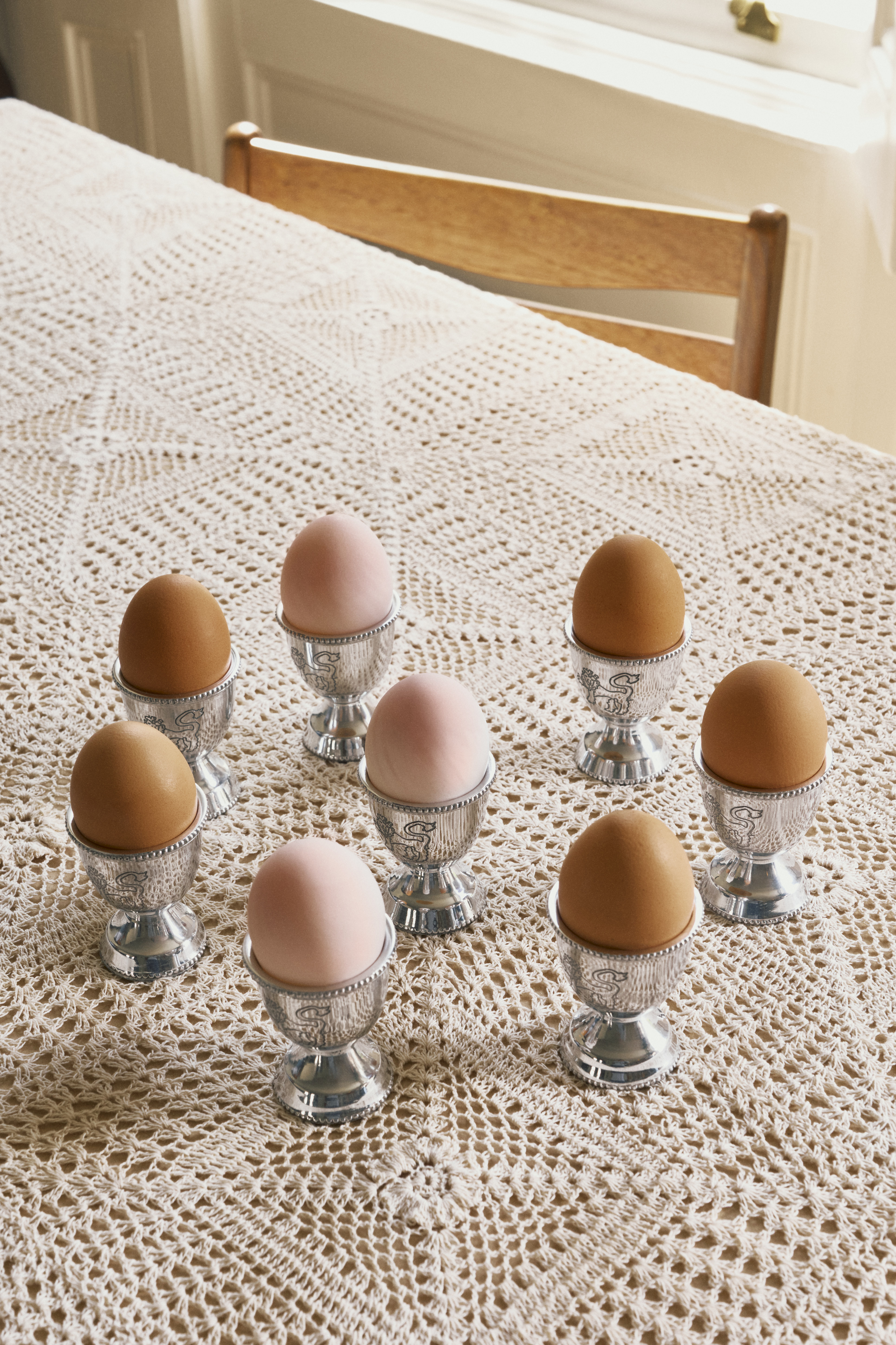 Close-up image of a table with a crochet tablecloth. There are eight silver egg cups on the table with eggs in them.