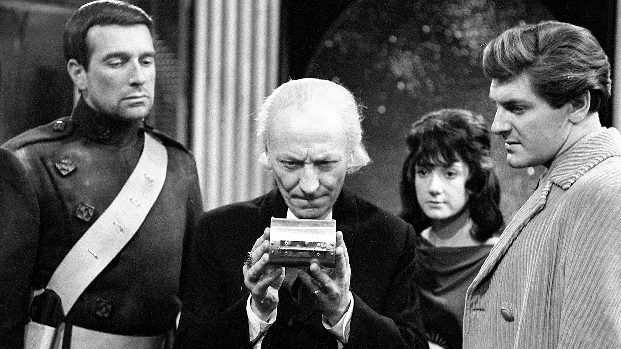 The first Doctor Who, with his companions in black and white, look intently at a small object. 