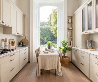 cream galley style kitchen with mix of wall and base units, large window at end and dining table in middle covered in cream cloth with wicker and wood chairs