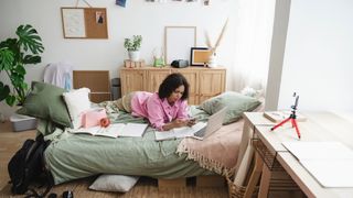 Woman lying on her front on a bed with her phone and laptop in a relatively cluttered room