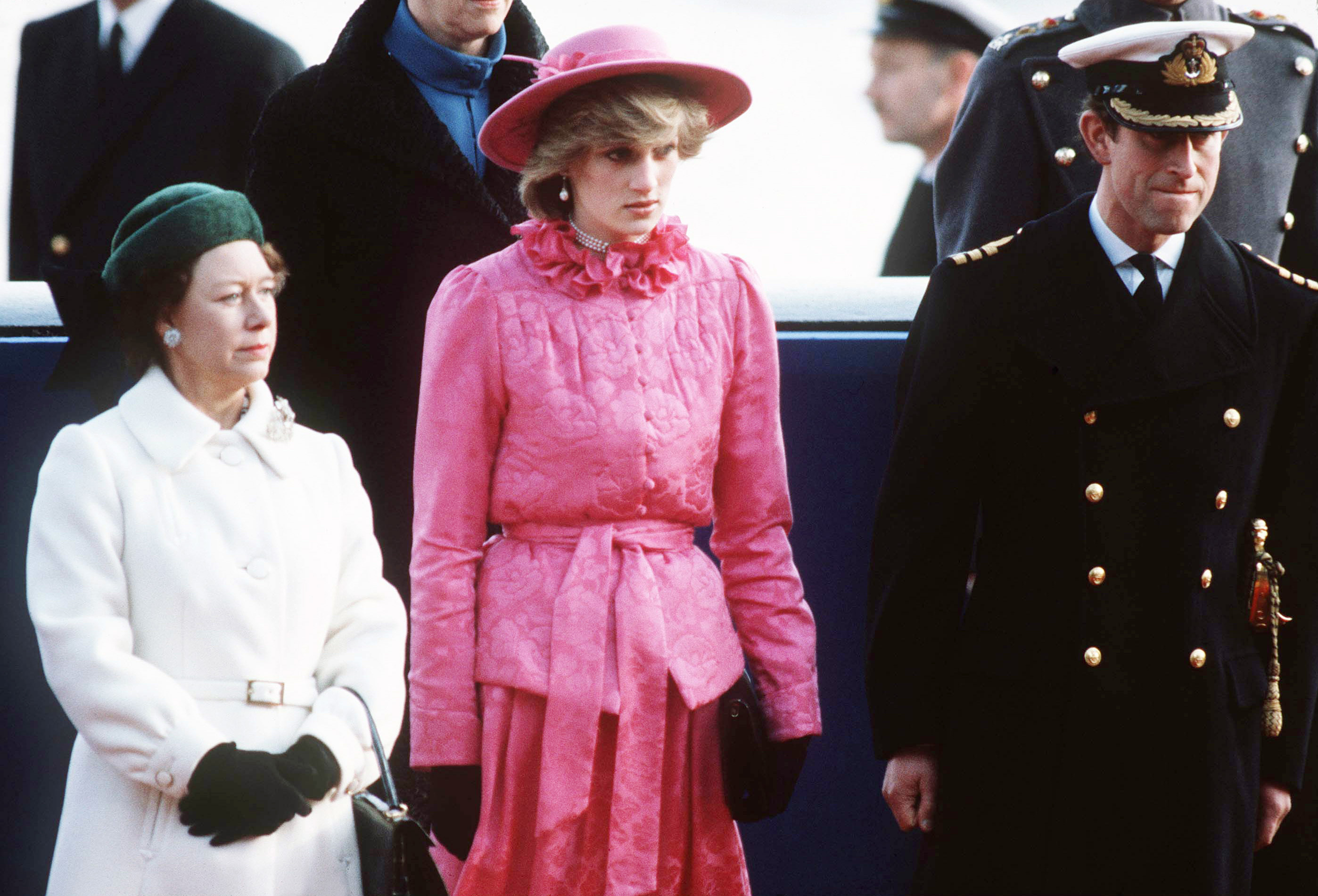Princess Margaret in a white coat standing next to Princess Diana wearing a pink outfit and Prince Charles in a military uniform