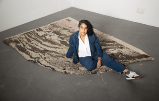 A woman dressed in a denim jacket and trousers, white shirt, and beige sneakers sits on a textile creations in the tones of beige, cream, and brown.