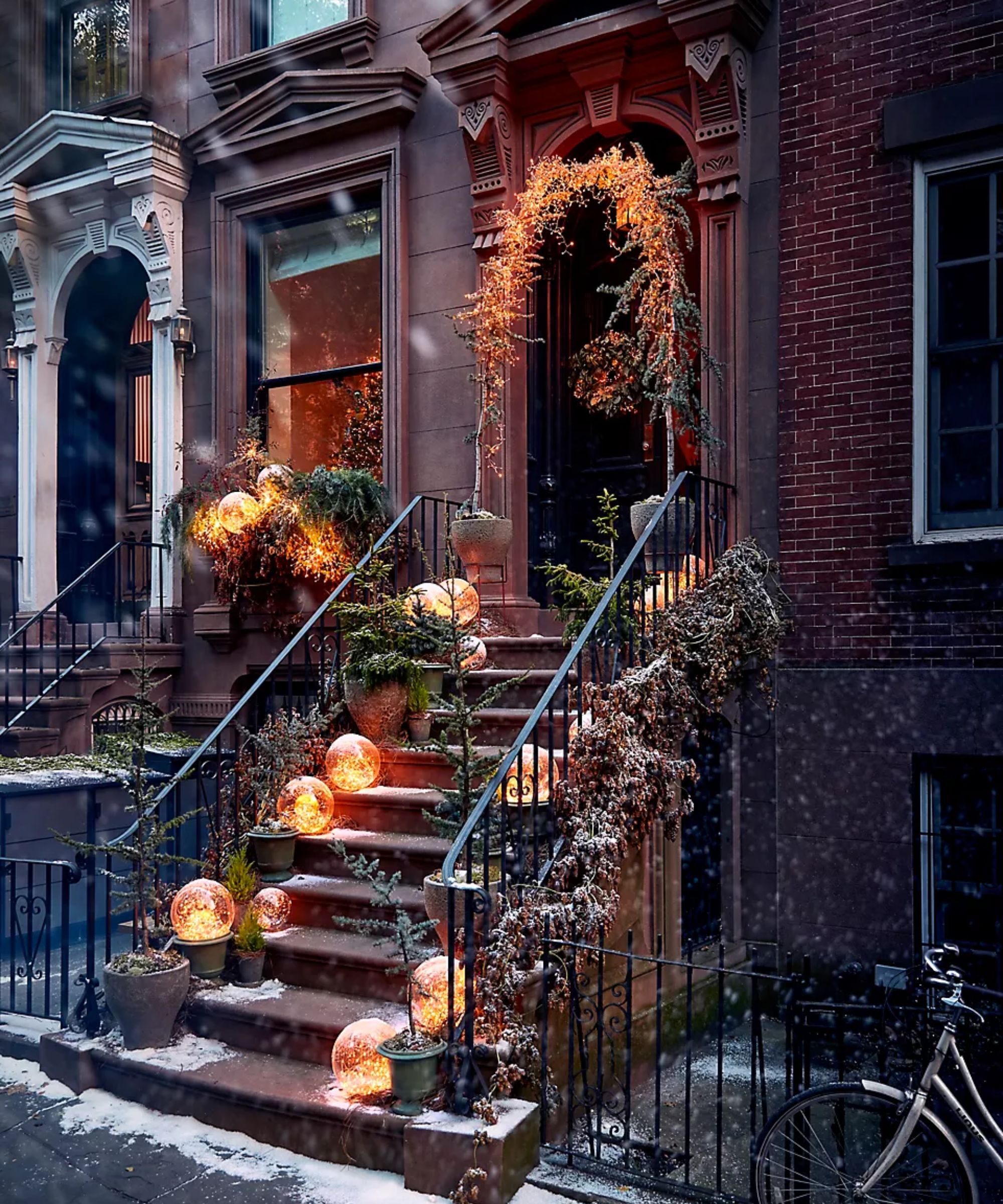 a brownstone town house with steps and door decorated for Christmas with a lit up garland and round christmas ball decor on the steps