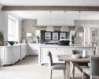 Bright, white kitchen with modern, country style, exposed stone wall, wooden beams on ceiling, white kitchen island and cabinets, marble countertops, artwork beside stove,