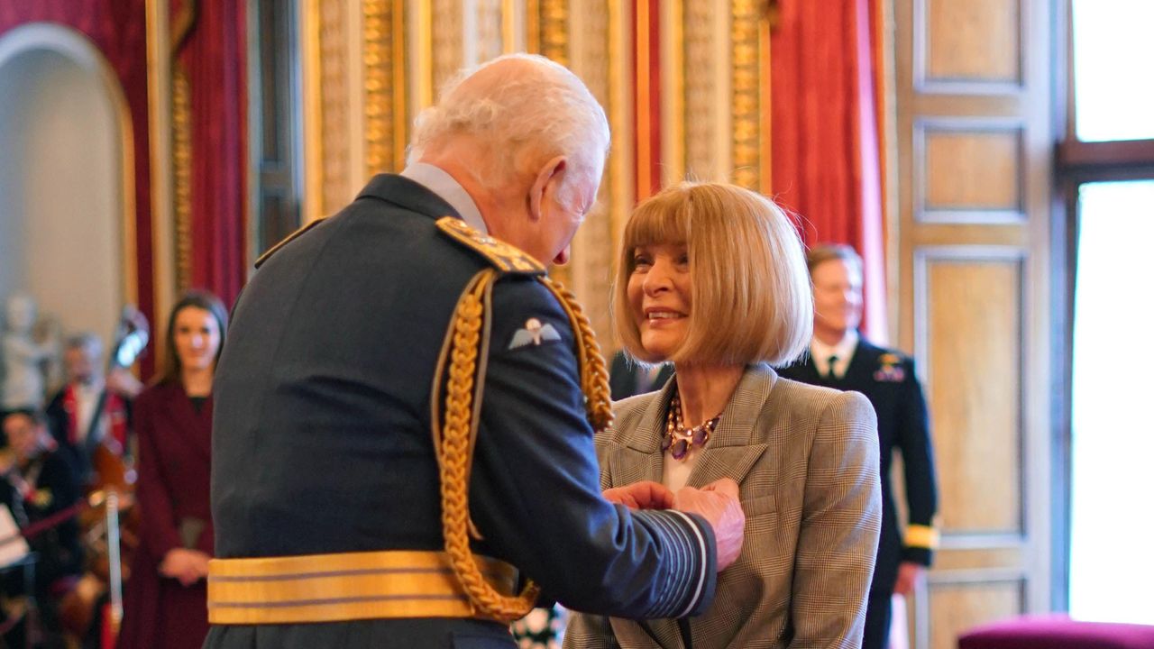 Anna Wintour wearing a gray blazer and smiling at King Charles as he pins a ribbon her jacket at Buckingham Palace