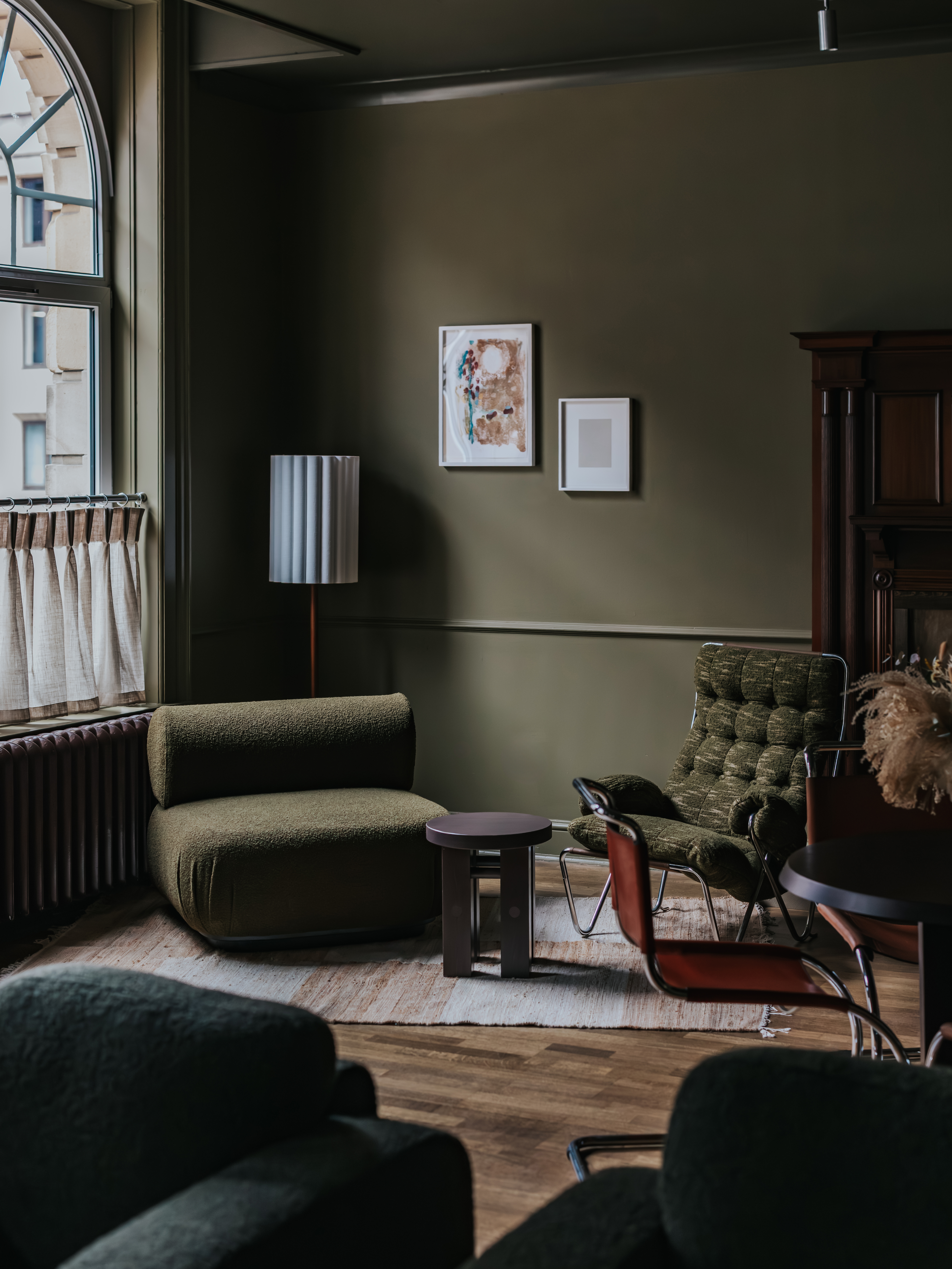 A dark, muted green-tinted hotel lounge with velvety chubby seating in similarly toned nuances, wall art, and wooden furniture.