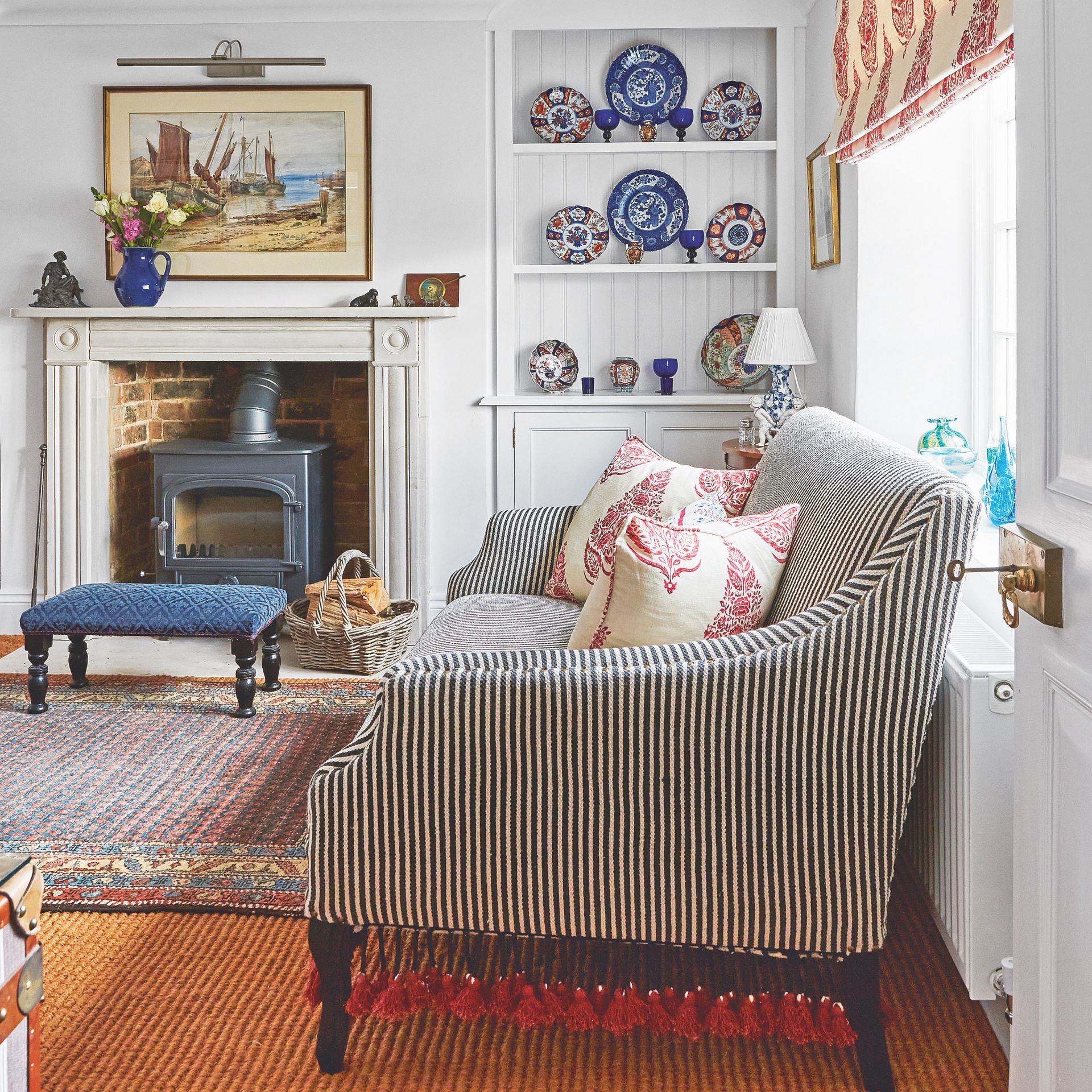 cottagecore decor ideas for living rooms, white living room with cottagecore style decor, plates in shelving, paisley blind, black and white stripe sofa, paisley cushions, log burner, artwork, jute carpet, rug