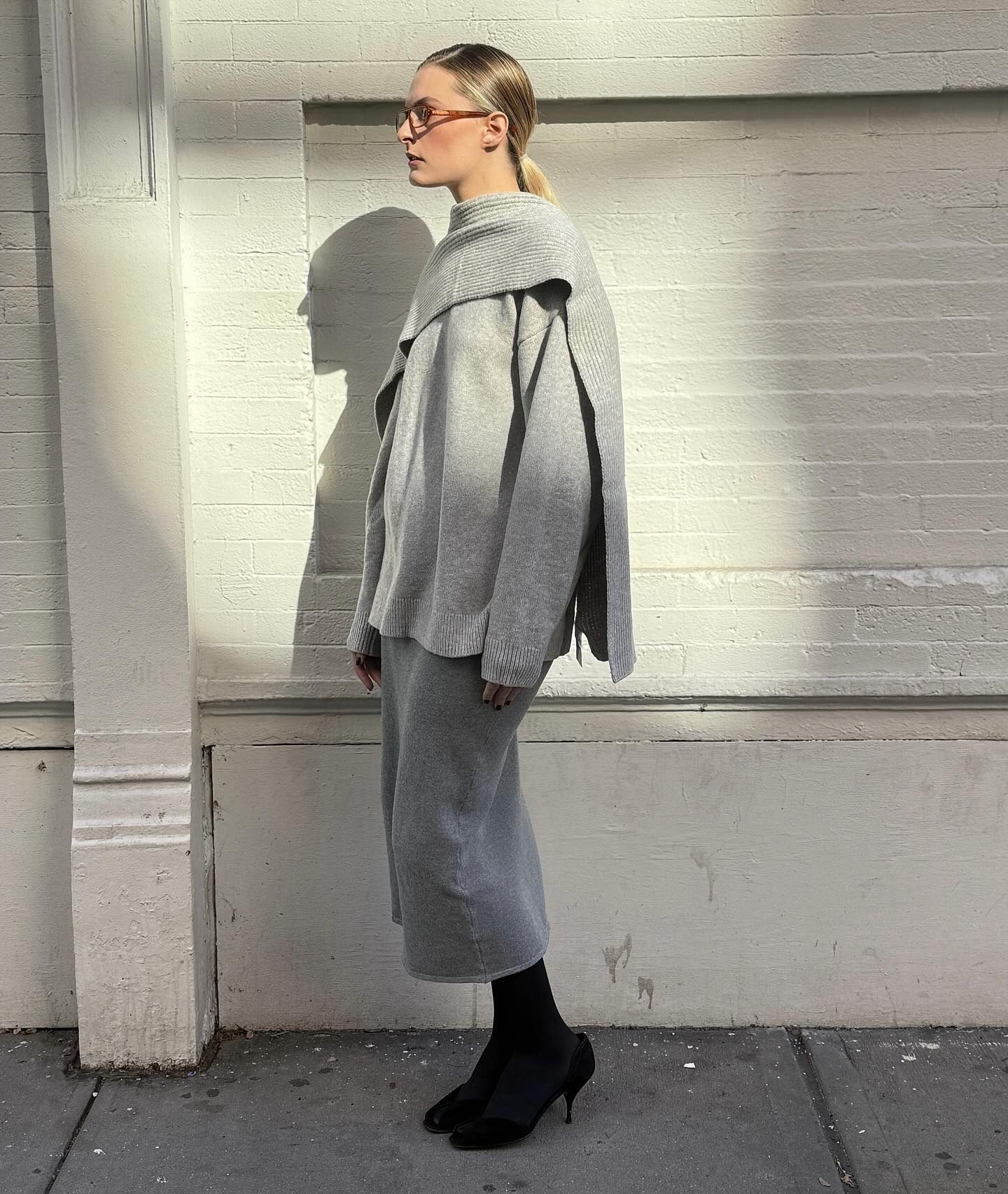 Influencer @christietyler poses outside in front of a beige brick wall wearing a grey cape jumper with a tonal grey skirt, thick black tights and black heels.