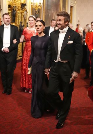 Victoria Beckham wearing a black floor length gown walking next to David Beckham wearing a tuxedo on a red carpet at Buckingham Palace