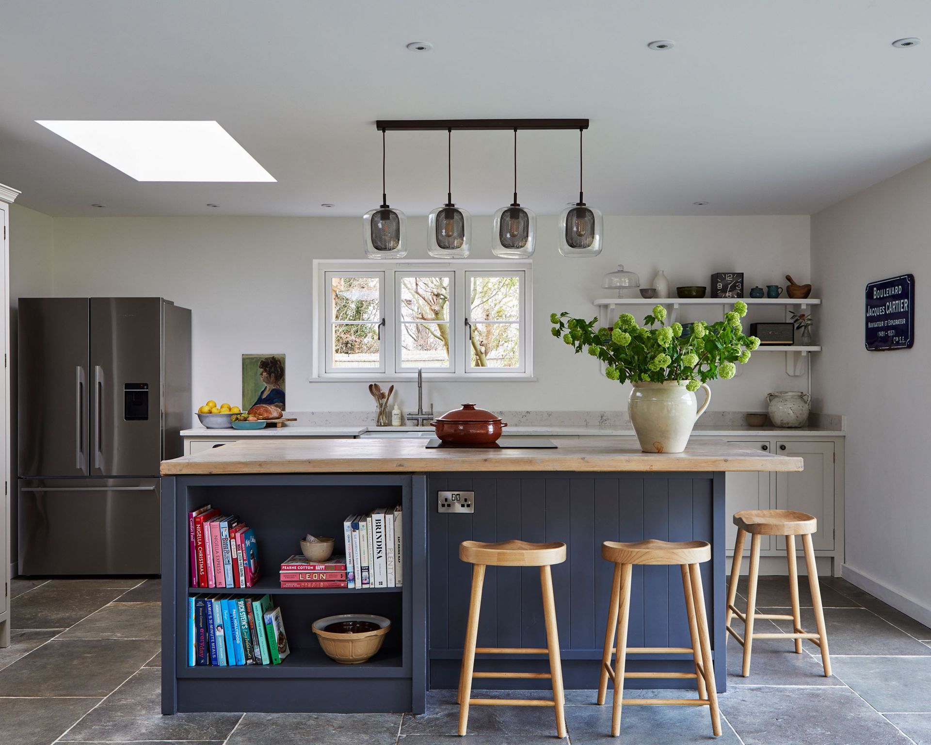 An open plan kitchen with dark blue cabinets on an island with wooden bar stools and a gray stone floor