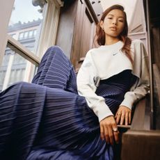 Woman wearing blue pleat skirt, white Nike jumper, and silver rings, sat in stairwell. 