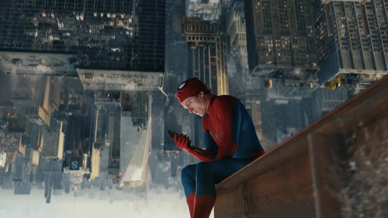 tom holland as spiderman lifting up his mask to look at a device while sitting on a building in spiderman brand new day