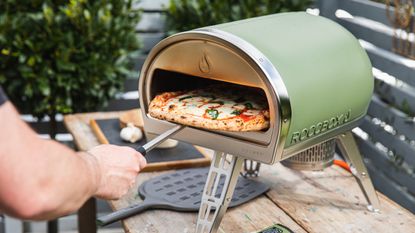 Person sliding a freshly cooked pizza out of a pale green pizza oven 