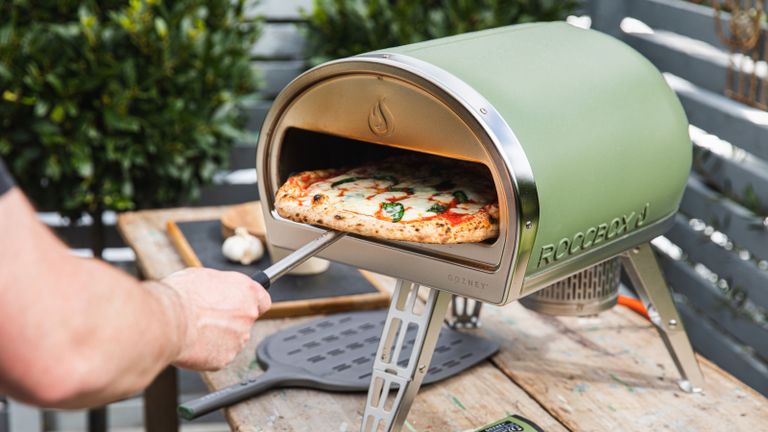 Person sliding a freshly cooked pizza out of a pale green pizza oven 