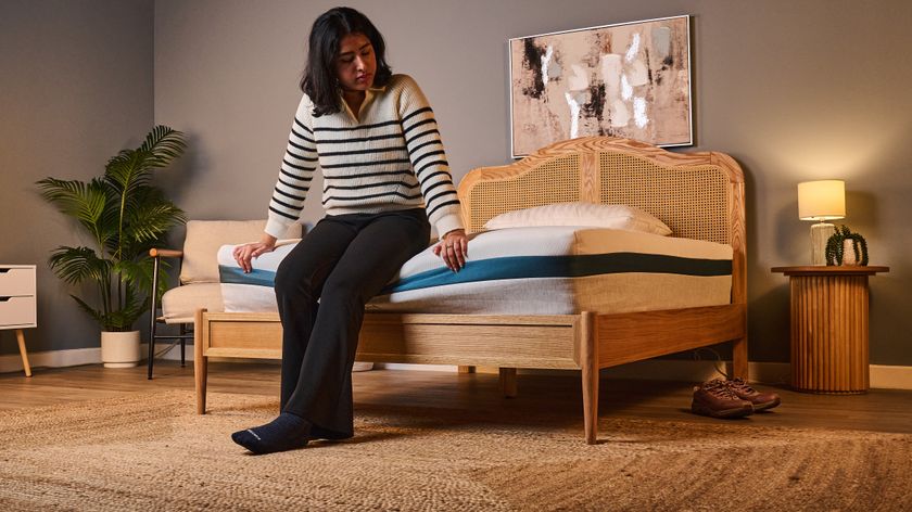 The image shows a woman sitting at the foot of the Helix Twilight mattress, which is placed on a wooden bed frame in a neutrally decorated bedroom