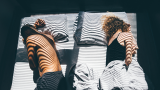 A couple sharing a bed, each one sleeping on their side facing the other way with light shining on them in stripes through a blind